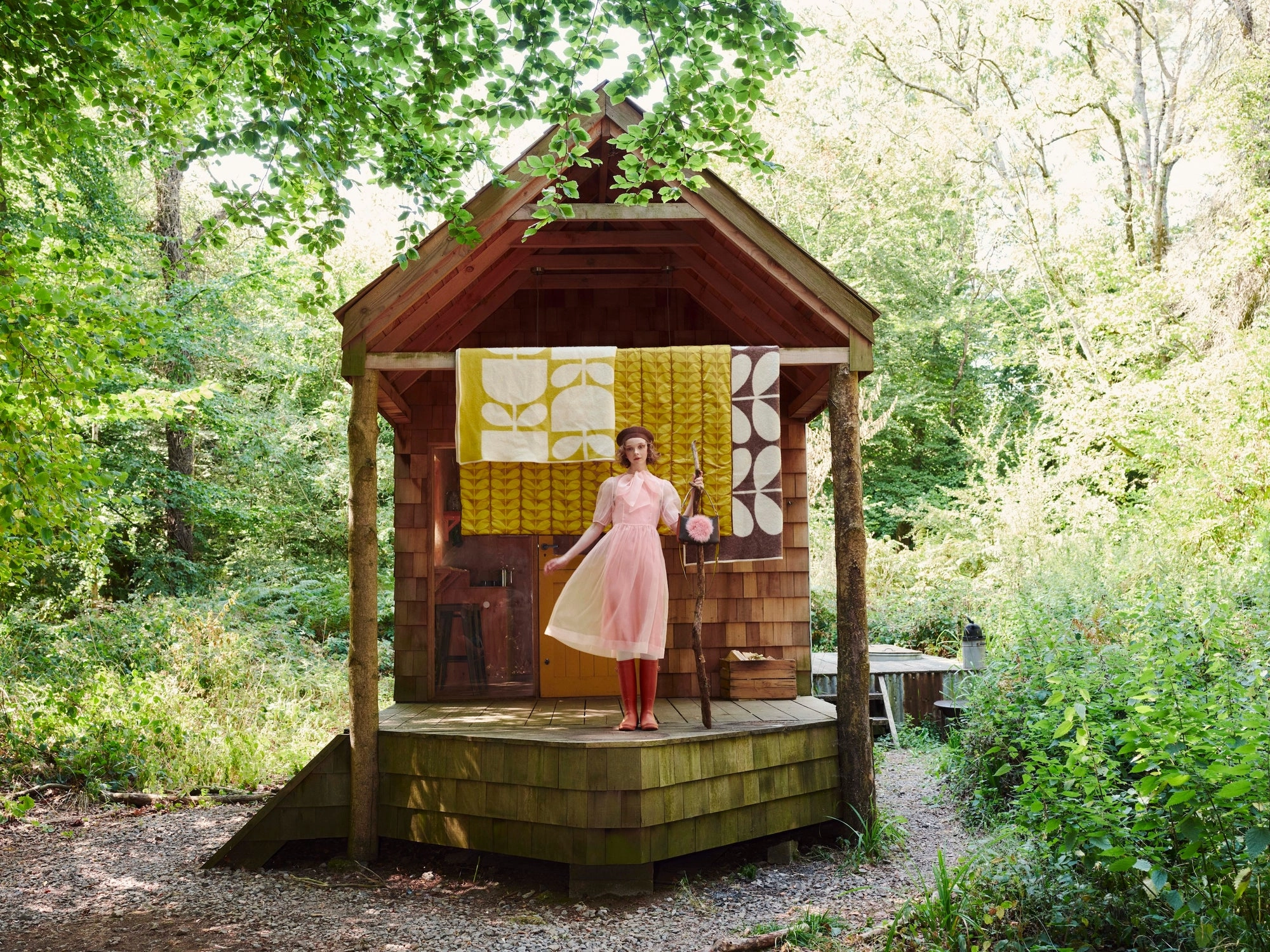 Orla Kiely AW26 look: a figure in a pink dress in front of a small wooden hut hung with yellow fabric